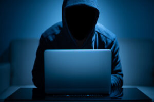 A shadowed person in a hoodie stealing sensitive information on a laptop. Prevent identity theft.