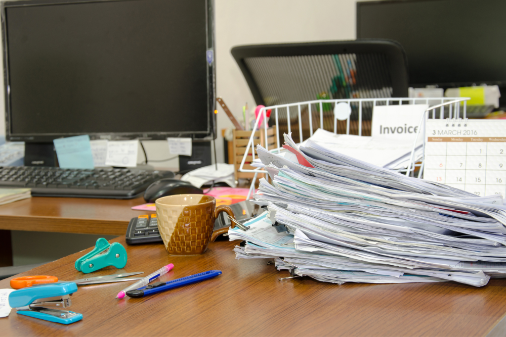 Disorganized office desk with a large pile of old invoices and paperwork, illustrating the need for outdated records shredding to maintain a clean, compliant workspace.